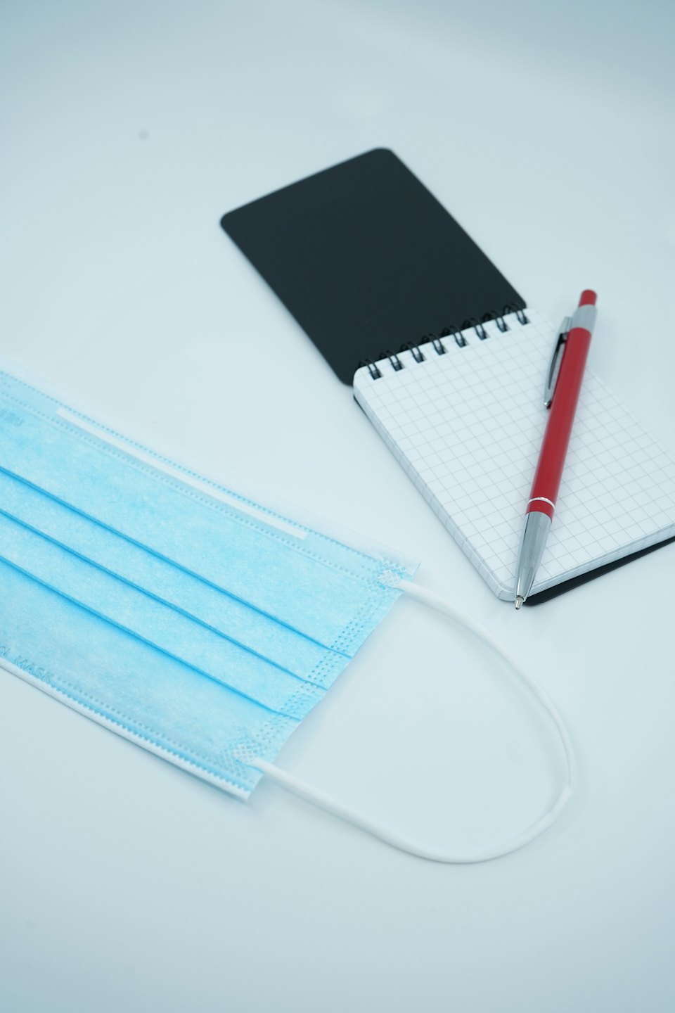 A blue medical face mask, notebook, and pen on a white surface.