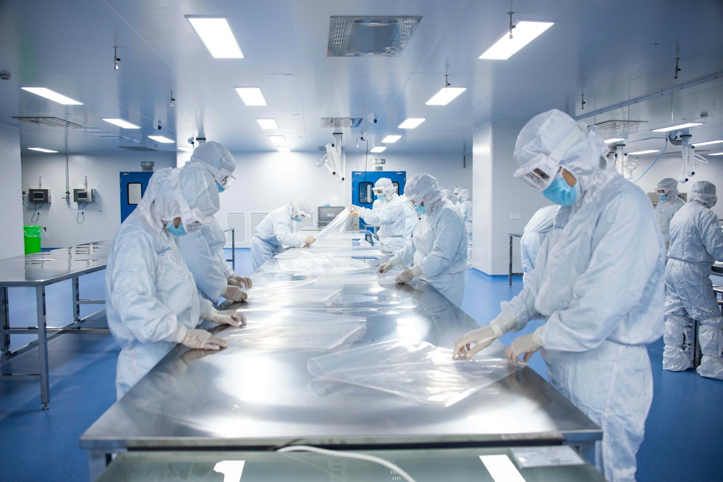 Workers in cleanroom suits handle plastic sheets on stainless steel tables.