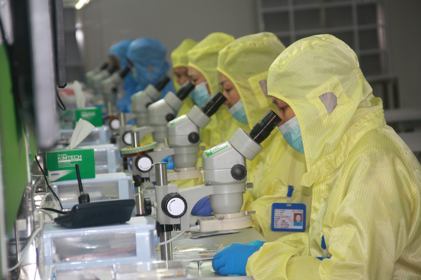 Workers in cleanroom suits inspecting items under microscopes.